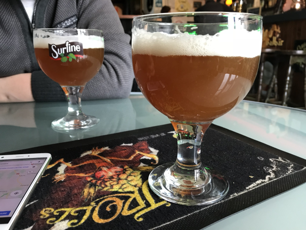 Zwei Gläser goldenen Bieres auf einem Tisch neben einem Handy, mit Menschen im Hintergrund und gerahmten Bildern an der Wand.