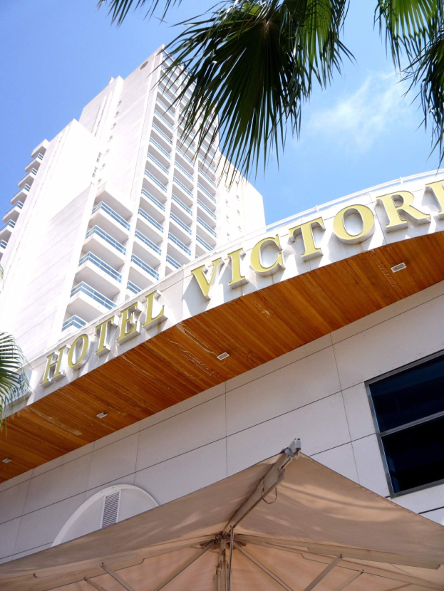 Das Bild zeigt das Hotel Victoria in Miami Beach, Florida, einen hohen Turm mit Glasfenstern und einer Namenstafel an der Wand, mit einem Zelt und einem Baum unten und weißen, flauschigen Wolken am Himmel.