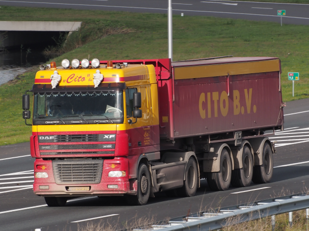 Ein roter und gelber Lkw fährt auf einer Autobahn, umgeben von einem Metallzaun, Gras, einem Pfahl, einem Schild, einer Brücke und einem Gewässer.