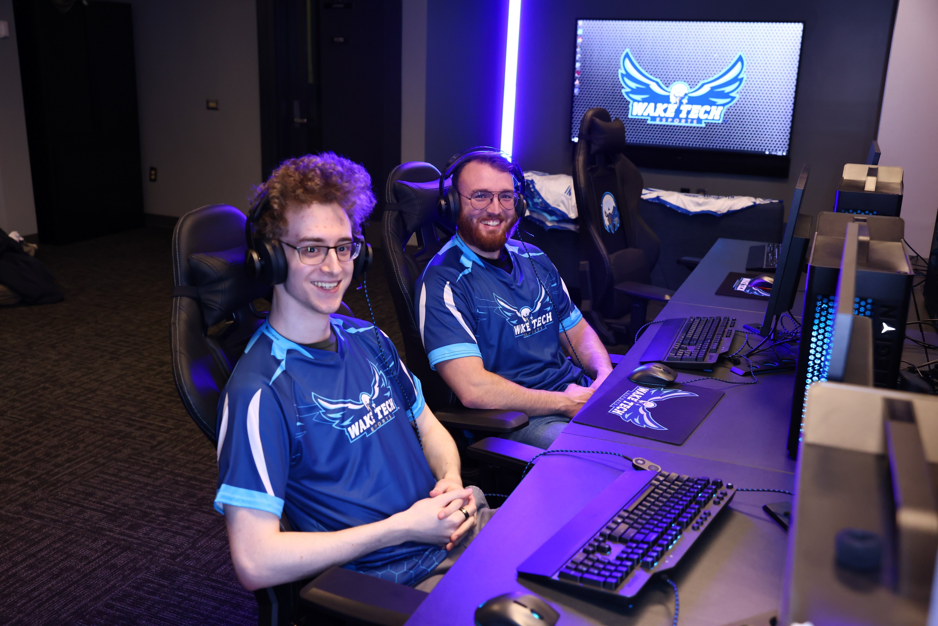 Zwei Männer in Headsets lächeln, während sie an einem Schreibtisch mit Computer-Monitoren, Tastaturen und anderem Equipment sitzen und scheinbar ein Spiel in einem Raum mit einem Bildschirm und Lichtern im Hintergrund spielen.