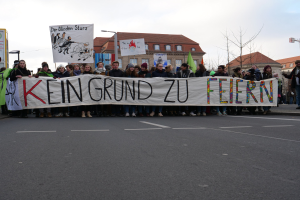 Demonstranten mit einem Transparent mit der Aufschrift 'Kein Grund zur Feier' gegen deutsche Sparmaßnahmen, mit Gebäuden, Bäumen und einem klaren Himmel im Hintergrund.
