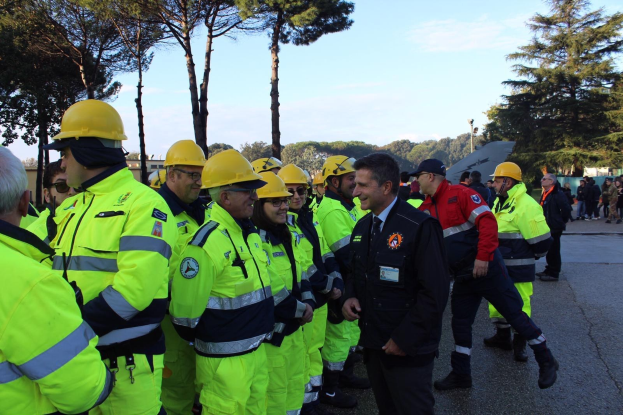 Gruppe von Menschen in gelben Westen und Helmen auf einer Straße neben Bäumen unter einem klaren blauen Himmel, einige tragen Mützen, was auf ein Einsatzteam hinweist.
