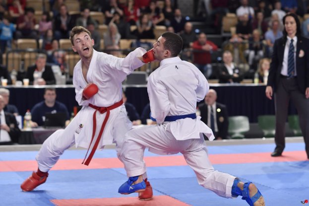 Zwei Männer in Karate-Ausrüstung im Judomatch, umgeben von Zuschauern auf Stühlen und Stehplätzen in der Nähe eines Tisches mit Flaschen und Gegenständen im Vordergrund.