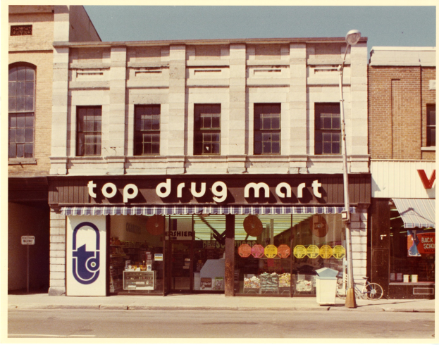 Straßensicht eines Top-Drogenmart-Geschäftsfronts an einer Straßenecke, mit einem Gebäude mit Fenstern und einer Schautafel, einem Straßenpfosten, einem geparkten Fahrrad, einem Müllcontainer und einem bewölkten Himmel.