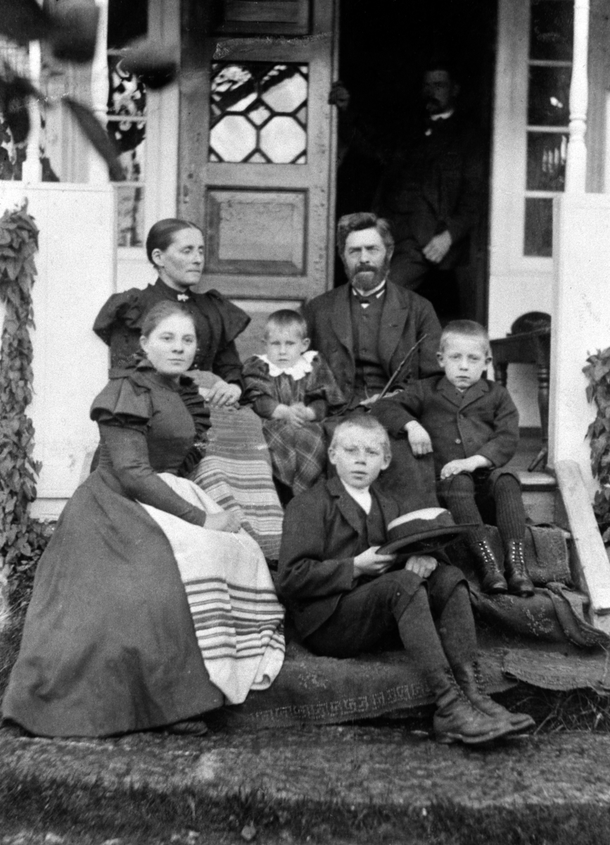 Ein Schwarz-Weiß-Foto einer Familie von fünf Personen, die auf den Stufen eines Hauses sitzen, mit einem Mann im Hintergrund.