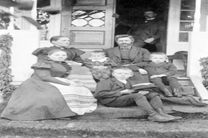 Ein Schwarz-Weiß-Foto einer Familie von fünf Personen, die auf den Stufen eines Hauses sitzen, mit einem Mann im Hintergrund.