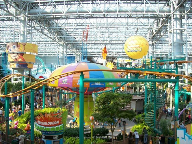 Großes Indoor-Wasserpark mit Achterbahn, umgeben von Menschen, Bäumen, Pflanzen und Deckenleuchten mit Stangen.