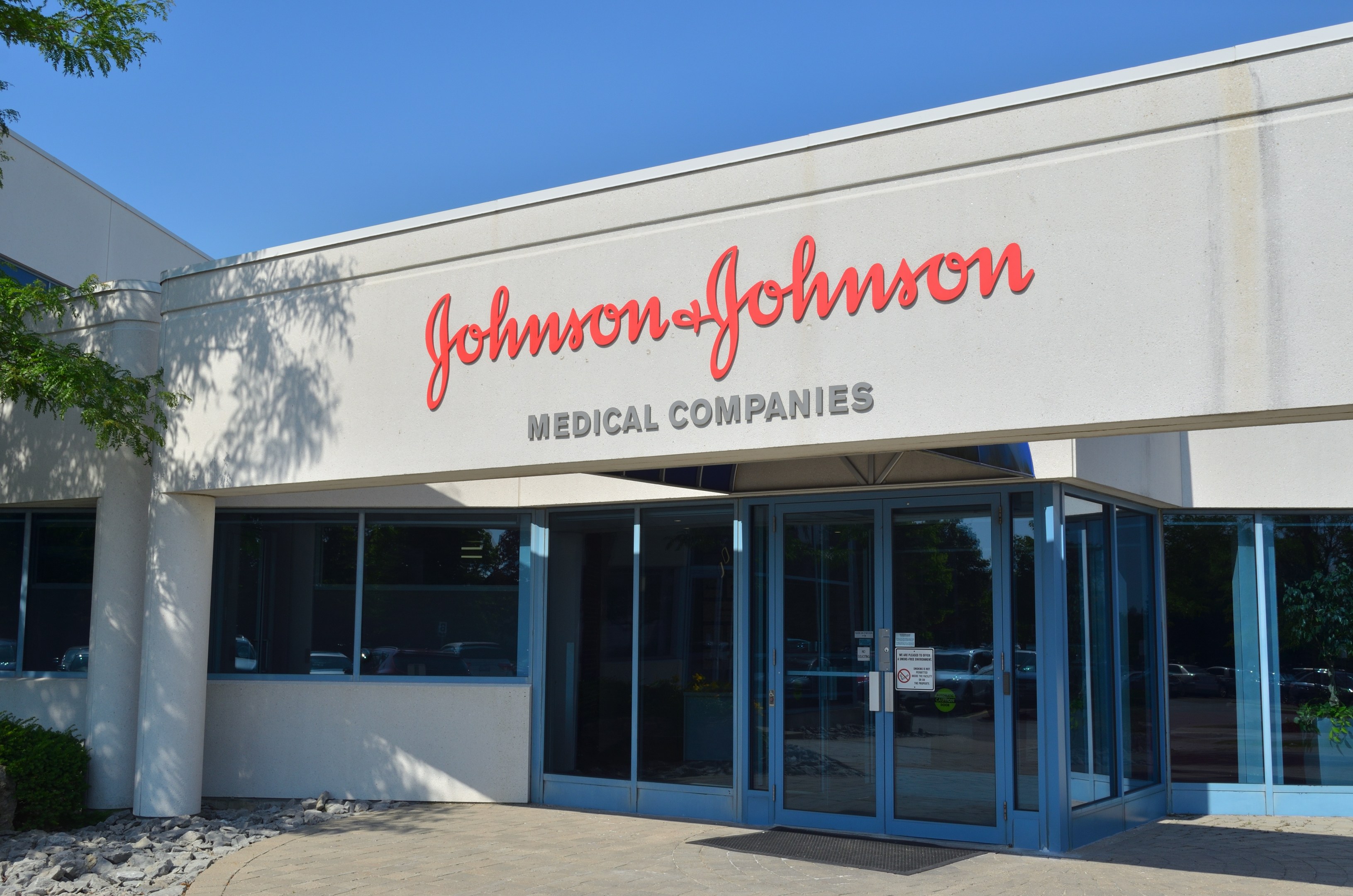 Außenansicht des Johnson & Johnson Medical Companies-Gebäudes mit Glaswänden, Text an der Fassade, Steinen, Pflanzen und einem Baum auf der linken Seite bei klarem blauem Himmel.