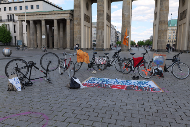 Eine Gruppe von Fahrr├Ądern, die vor dem Brandenburgertor in Berlin, Deutschland, geparkt sind, mit Taschen und einer Fahne in der N├Ąhe, vor Geb├Ąuden, B├Ąumen, Masten, Flaggen und einem klaren blauen Himmel.