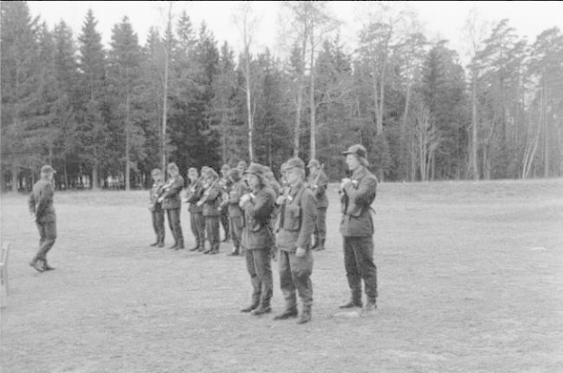 Schwarz-weißes Bild einer Gruppe von Männern in Militäruniformen mit Mützen und Gewehren, die in einem Feld mit Bäumen und einem klaren Himmel im Hintergrund stehen, wahrscheinlich während einer Übung.