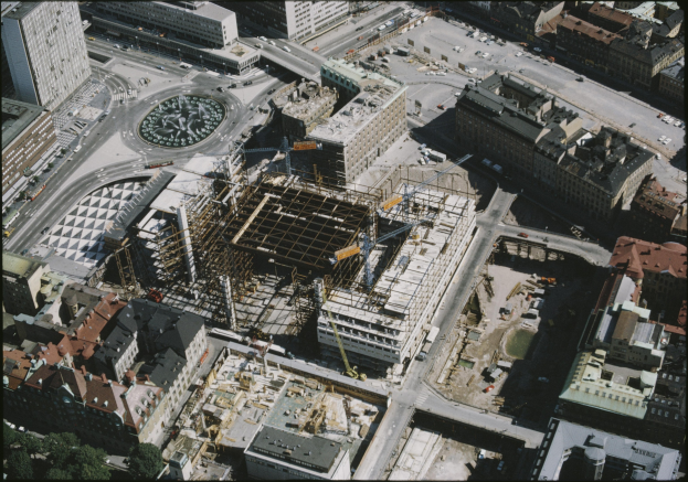 Luftaufnahme einer Stadt mit einem großen Gebäude in der Mitte, umgeben von anderen Gebäuden, Straßen mit Fahrzeugen und Bäumen, mit Text unten.