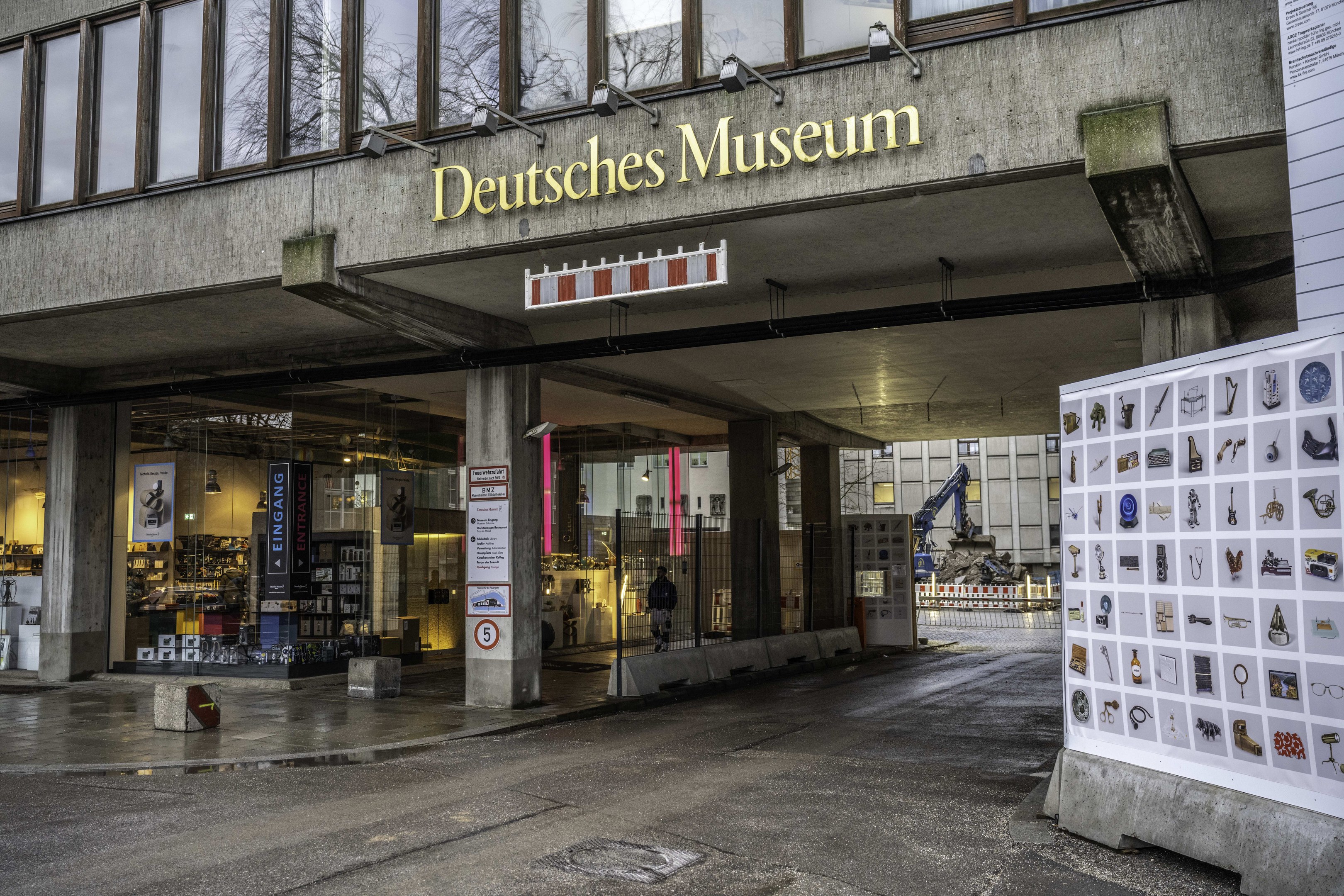Großes modernes Gebäude mit Glasfenstern und Säulen, beschriftet mit "Deutsches Museum", mit einer informativen Schautafel auf der rechten Seite, vor dem Hintergrund anderer Gebäude, Bäume und Himmel.