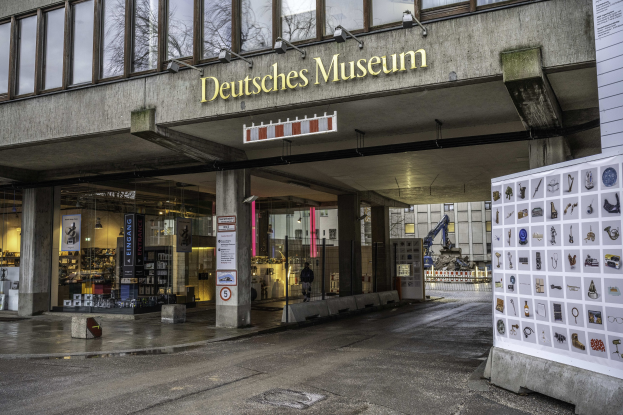 Großes modernes Gebäude mit Glasfenstern und Säulen, beschriftet mit "Deutsches Museum", mit einer informativen Schautafel auf der rechten Seite, vor dem Hintergrund anderer Gebäude, Bäume und Himmel.