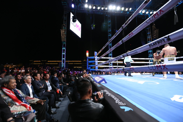 Schwarz-weißes Foto von zwei Boxern in einem Boxring mit Helmen und Handschuhen, umgeben von Seilen unter beleuchteten Lichtern, mit 'Olympian Magazine'-Text oben und unten.