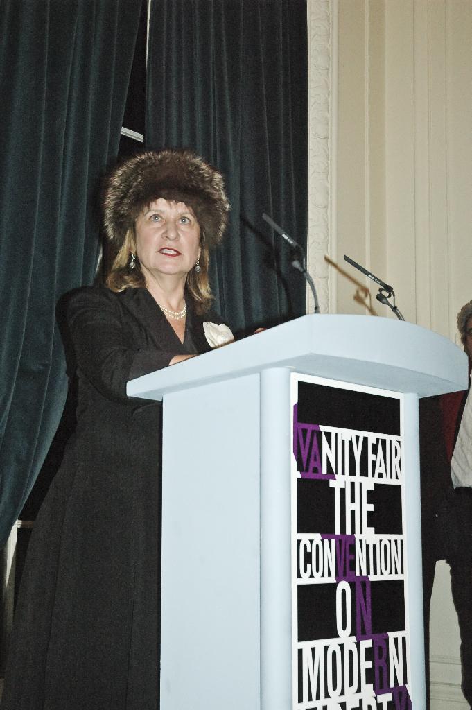 Eine Person steht an einem Rednerpult mit Text darauf, spricht in ein Mikrofon, mit mehreren Personen und Vorh√§ngen im Hintergrund.