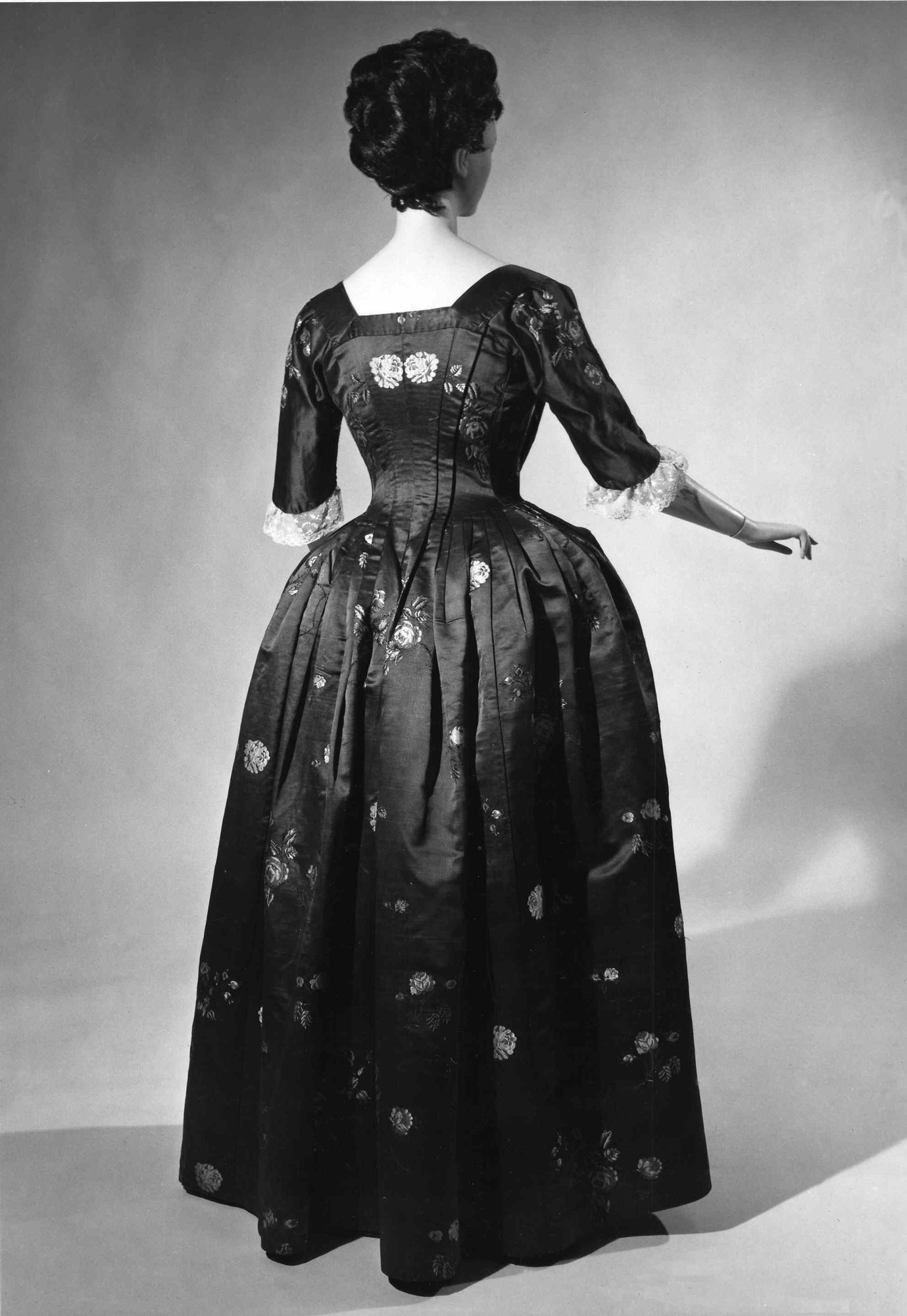 Eine Frau in einem Schwarz-Weiß-Foto aus den 1950er Jahren in einem langen, eng anliegenden Kleid mit einem luxuriösen Stoff, gekreuzten Händen, klassischem Hochsteckdutt, schwarzen High Heels und dezentem Make-up.