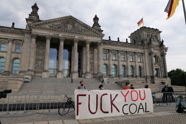 Demonstranten mit einem Transparent mit der Aufschrift "Fuck You Coal" vor dem Reichstagsgebäude in Berlin, Deutschland, mit den architektonischen Details des Gebäudes, einem Fahrrad in der Nähe, Bäumen, einer Fahne an einem Fahnenmast und einem bewölkten Himmel.