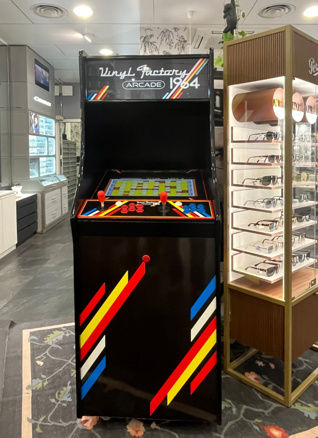 Ein Geschäft mit einem zentralen Retro-Arcade-Automaten, einem Regal mit Gläsern auf der rechten Seite und Hintergrund-Elementen wie Schränken, einem Fernseher und anderen Gegenständen, sowie Deckenleuchten, die eine Retro-Atmosphäre schaffen.