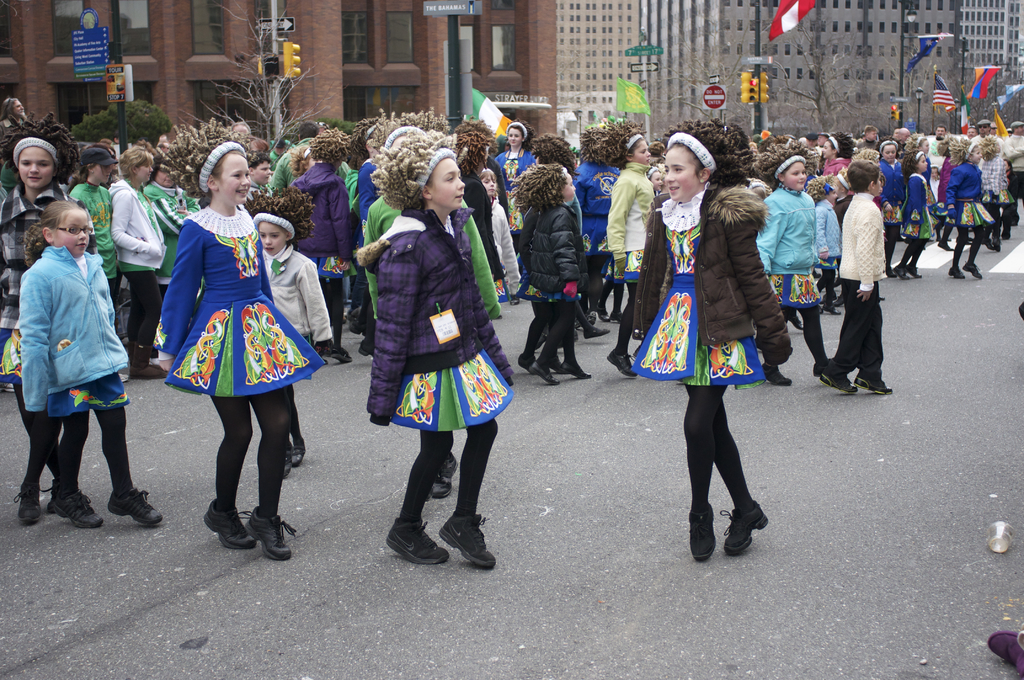 Eine Gruppe von Mädchen in Kleidern mit Jacken und Pullover, die auf einer Straße tanzen, mit einem roten Gebäude und einem gelben Signalmast dahinter und Gebäuden und Fahnen auf Strommasten im Hintergrund.
