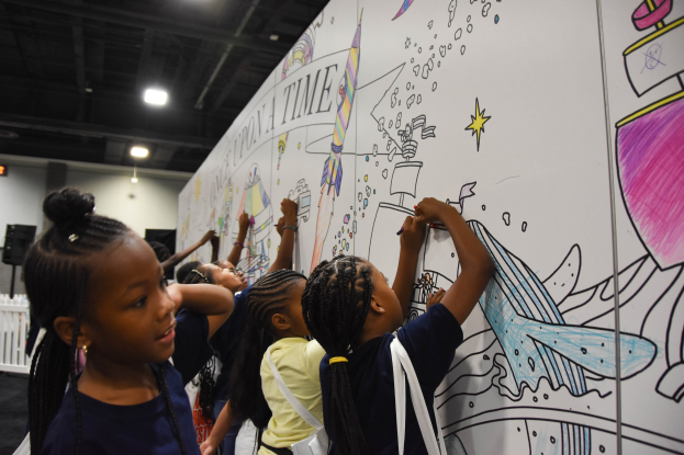 Eine Gruppe von Kindern malt auf einer großen Whiteboard mit Markern während einer Kunstausstellung, umgeben von einem Zaun, Deckenleuchten und Lautsprechern.