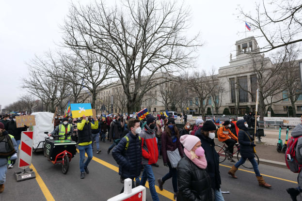 Eine große Gruppe von Menschen nimmt an einer Protestdemo in Washington, D.C. am 21. Januar 2020 teil, die eine Straße entlangmarschiert, Schilder und Banner schwingt und Fahrräder fährt, vor einer Kulisse aus Schautafeln, Bäumen und einem klaren blauen Himmel.