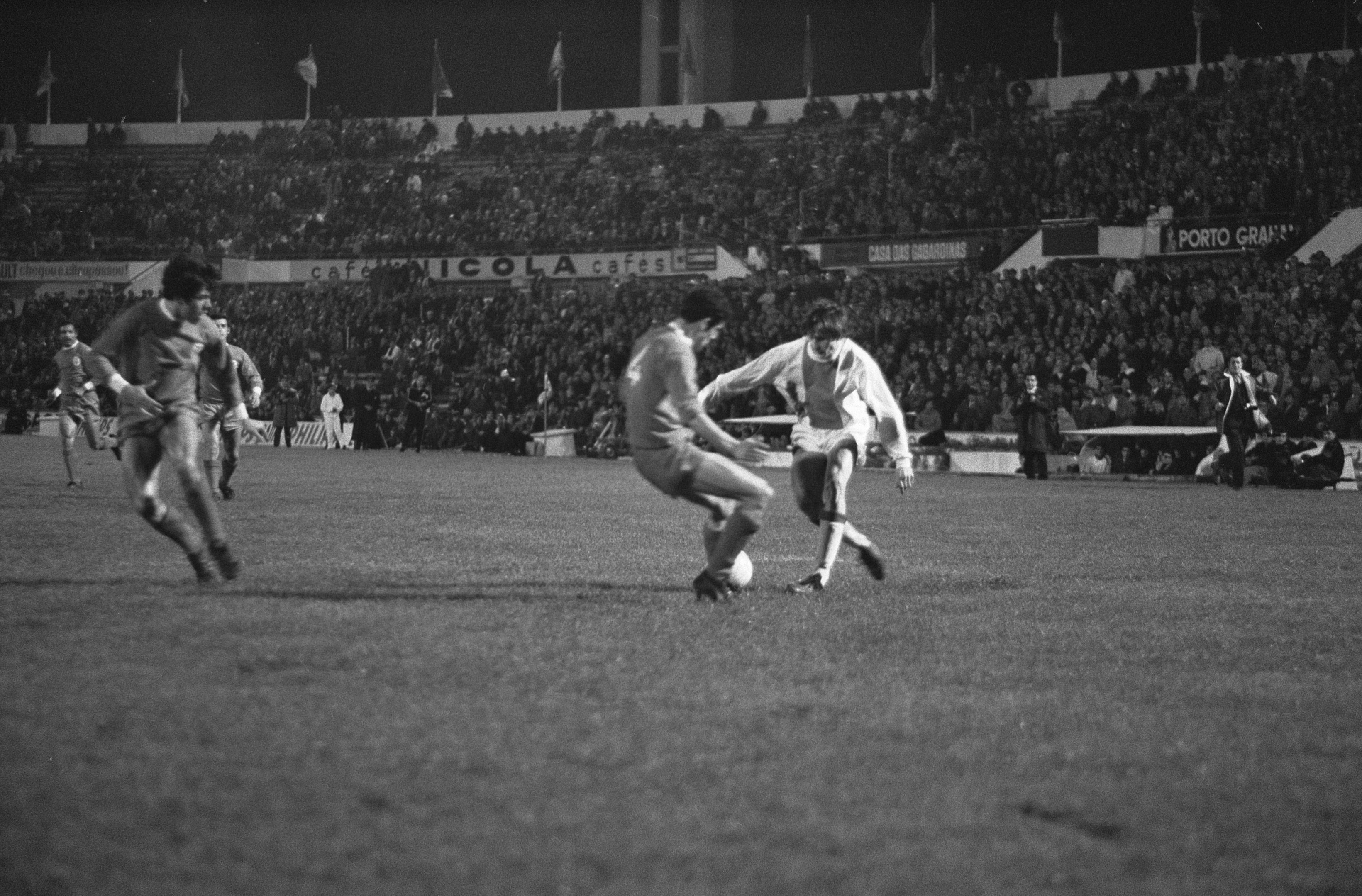Schwarzes und weisses Bild von Männern, die Fussball auf einem Feld in einem Stadion mit Zuschauern in den Rängen und Fahnen im Hintergrund spielen.