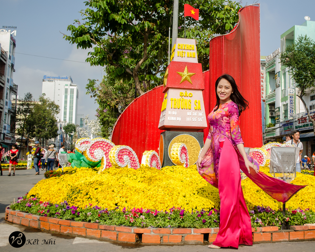 Eine Gruppe von Menschen mit einer lachenden Frau in der Mitte, umgeben von Ziegeln, Blumen, einer Fahne auf einem Turm, mit Gebäuden, Bäumen und Plakaten im Hintergrund.