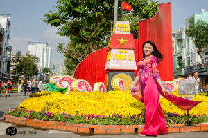 Eine Gruppe von Menschen mit einer lachenden Frau in der Mitte, umgeben von Ziegeln, Blumen, einer Fahne auf einem Turm, mit Gebäuden, Bäumen und Plakaten im Hintergrund.