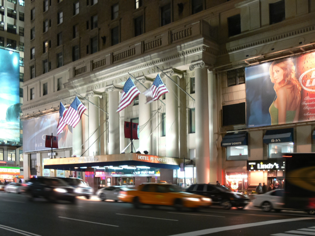 Belebte Stadtstraße bei Nacht mit fahrenden Fahrzeugen, einem Gebäude mit Fenstern, Säulen und Fahnenmasten im Hintergrund, beleuchteten Schildern mit Text und einem Bild einer Frau und Straßenbeleuchtung.
