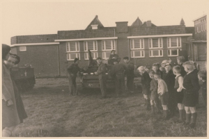 Eine Gruppe von Kindern steht um ein militärisches Fahrzeug vor einem Gebäude mit Fenstern, umgeben von Gras und einem klaren Himmel, in Schwarz-Weiß.