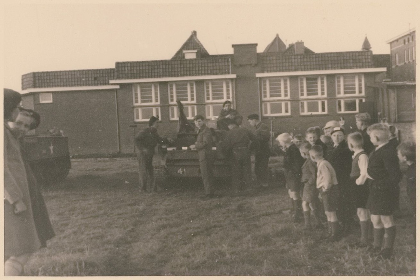 Eine Gruppe von Kindern steht um ein militärisches Fahrzeug vor einem Gebäude mit Fenstern, umgeben von Gras und einem klaren Himmel, in Schwarz-Weiß.
