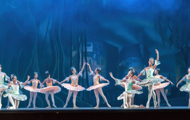 Eine Gruppe von Balletttänzerinnen in farbenfrohen Kostümen führt eine synchronisierte Choreographie auf der Bühne des Royal Opera House auf, vor einem blauen Hintergrund.