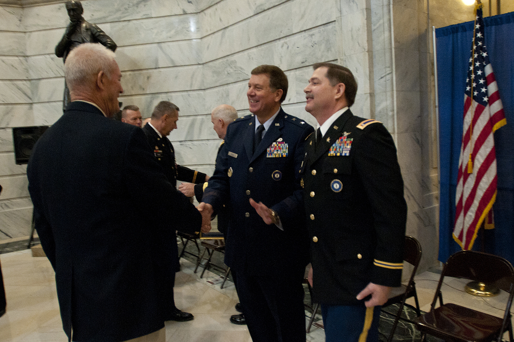 Menschen schütteln sich die Hände in einem Raum mit Stühlen, einer Statue neben einer Wand, einer Fahne und einem Vorhang auf der rechten Seite.
