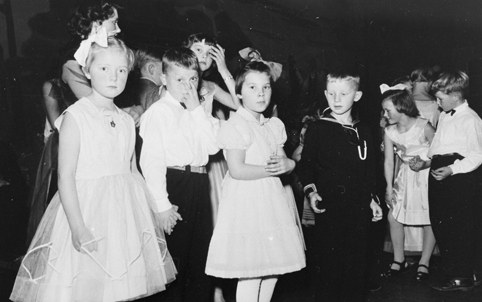 Schwarzes und weißes Foto einer Gruppe von Kindern, die auf einer Tanzfläche stehen, lächeln und glücklich aussehen, mit einer Wand im Hintergrund.