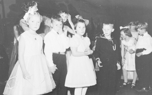 Schwarzes und weißes Foto einer Gruppe von Kindern, die auf einer Tanzfläche stehen, lächeln und glücklich aussehen, mit einer Wand im Hintergrund.