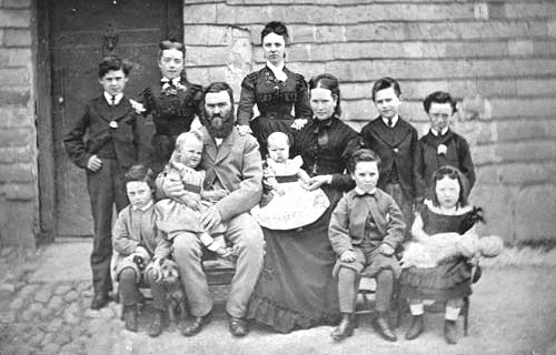 Schwarzes und weißes Foto einer Familie, die vor einem Haus posiert, wobei einige auf einer Bank sitzen und andere stehen, eine Wand und eine Tür sind im Hintergrund sichtbar.