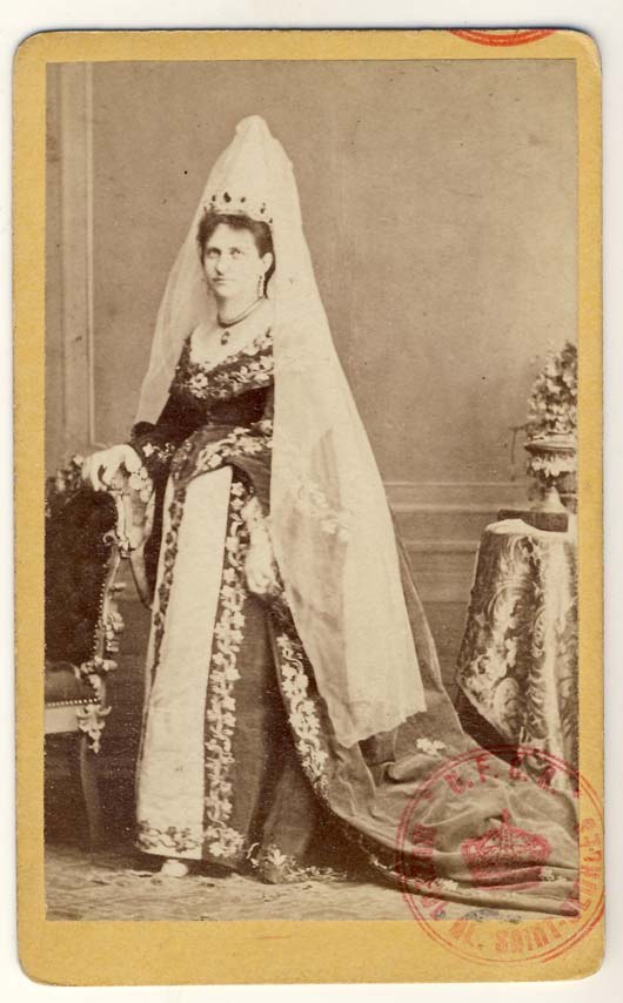 Ein altes Foto einer Frau in einem traditionellen russischen Hochzeitskleid, die auf einem Stuhl sitzt und einen Blumenstrauß hält, mit einem Tisch und einer Vase auf der rechten Seite und Text und einem Stempel unten.