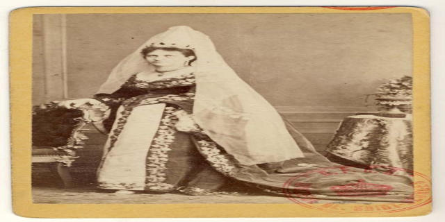 Ein altes Foto einer Frau in einem traditionellen russischen Hochzeitskleid, die auf einem Stuhl sitzt und einen Blumenstrauß hält, mit einem Tisch und einer Vase auf der rechten Seite und Text und einem Stempel unten.