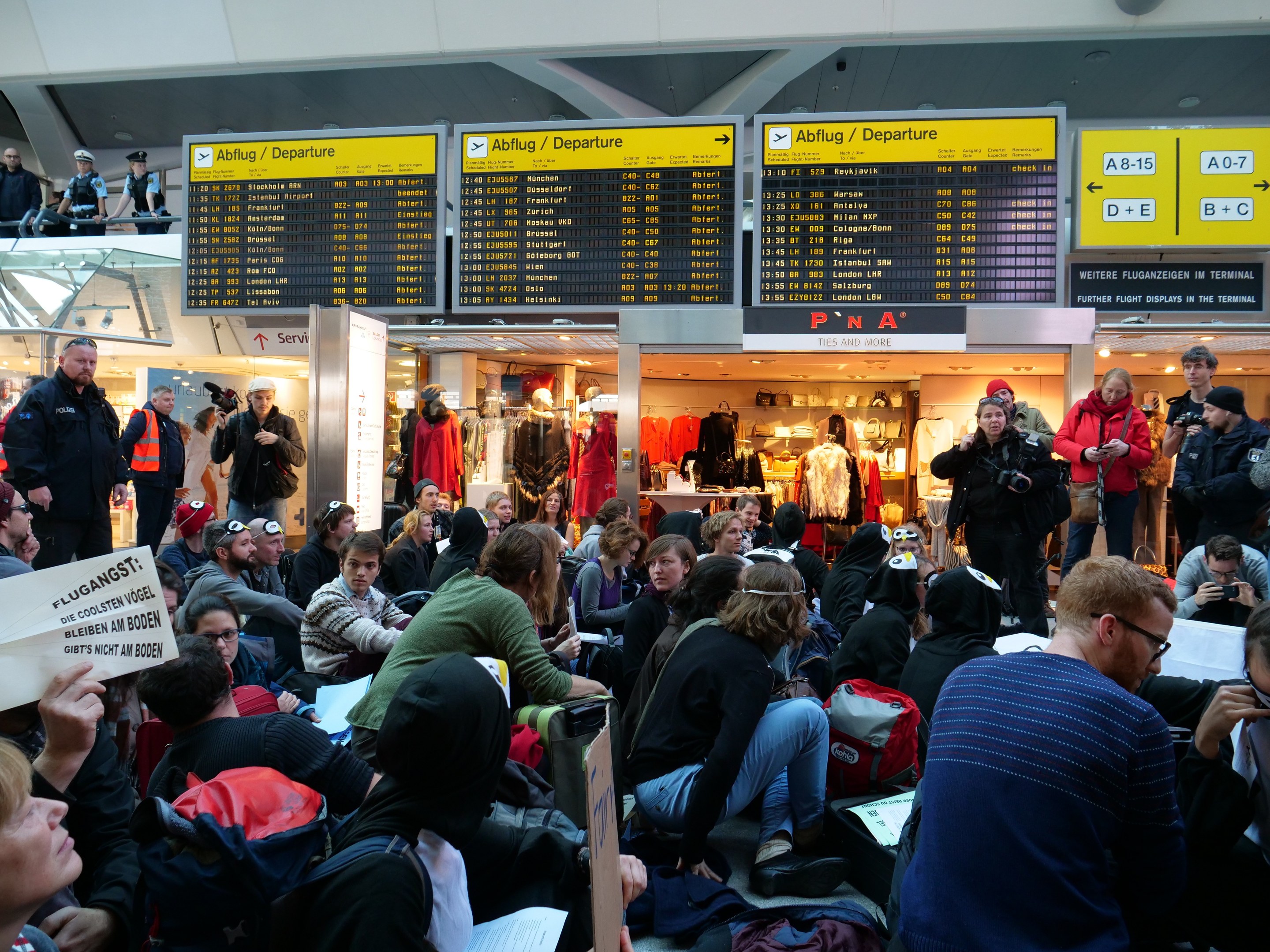 Eine große Gruppe von Menschen in einem Flughafen, einige sitzen mit Taschen und Papieren, andere stehen, mit Texttafeln, Schaufensterpuppen in Kleidern und Deckenleuchten, was auf eine Demonstration hindeutet.