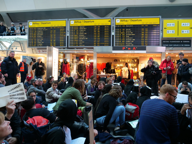 Eine große Gruppe von Menschen in einem Flughafen, einige sitzen mit Taschen und Papieren, andere stehen, mit Texttafeln, Schaufensterpuppen in Kleidern und Deckenleuchten, was auf eine Demonstration hindeutet.