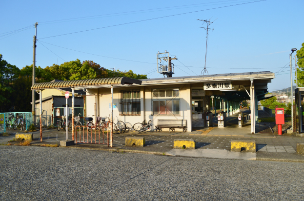Ein Bahnhof mit vor ihm geparkten Fahrrädern, eine Straße unten und Häuser, Bäume, Pfosten, Drähte und einen klaren blauen Himmel im Hintergrund.