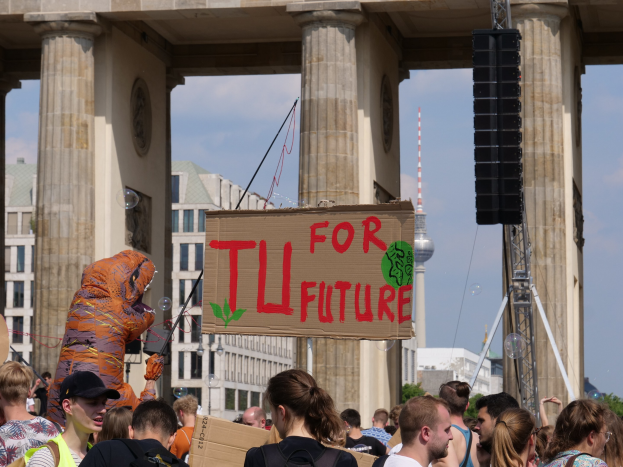 Eine Gruppe von Menschen vor dem Brandenburger Tor in Berlin, Deutschland, mit einer Person in Kostüm, die ein Schild hält, umgeben von Gebäuden, Bäumen und einem Turm unter einem bewölkten Himmel.