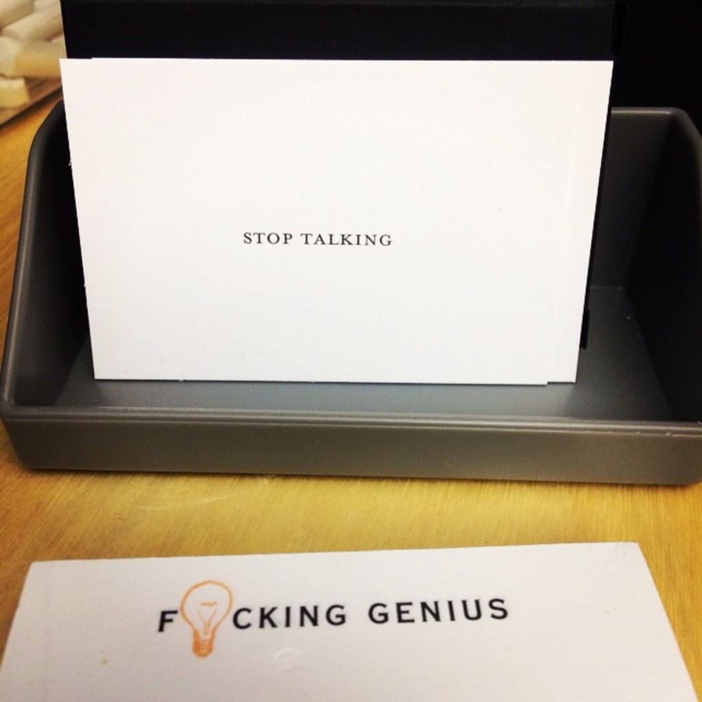 Ein Drucker-Karton mit einem Blatt Papier darauf, das den Text 'Hör auf zu reden' anzeigt, und einem weiteren Blatt auf dem Tisch, das den Text 'Verdammter Genius' anzeigt.