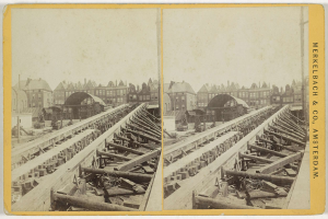 Schwarz-weißes Foto einer Zugschiene mit Gebäuden im Hintergrund, Text 'Merkelbach & Co, Co. Amsterdam' auf der rechten Seite und verstreute hölzerne Gegenstände im Vordergrund.