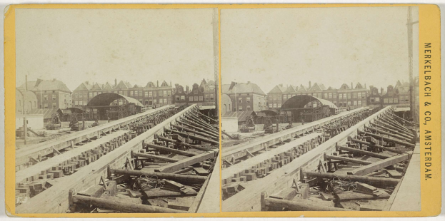 Schwarz-weißes Foto einer Zugschiene mit Gebäuden im Hintergrund, Text 'Merkelbach & Co, Co. Amsterdam' auf der rechten Seite und verstreute hölzerne Gegenstände im Vordergrund.