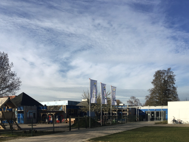 Großes Gebäude mit einem grasigen Bereich davor, umgeben von einem Metallzaun, mit einem Weg, Bäumen, Pfosten, Schildern, einem geparkten Fahrrad, nahen Häusern und einem bewölkten Himmel, identifiziert als der neue Spielbereich für Kinder an der Schule.