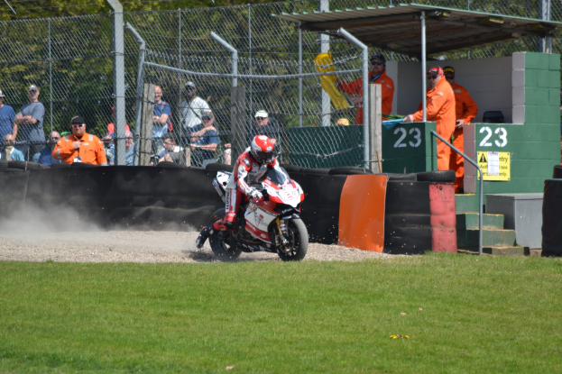 Eine Person in Helm und Handschuhen fährt ein Motorrad über eine grüne Wiese, mit Zuschauern, Absperrungen, Treppen und einem nummerierten Schuppen im Hintergrund.