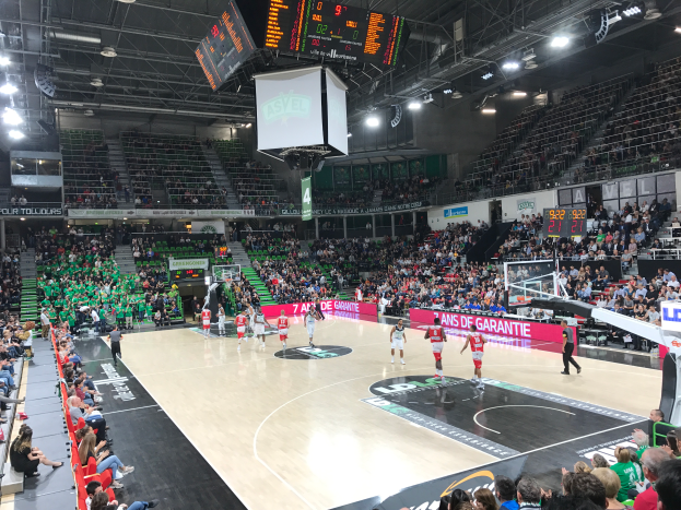 Ein Basketballspiel in einer großen Arena mit Zuschauern in den Rängen, Anzeigetafeln mit "Asvel vs Galatasaray" und einer beleuchteten Decke mit einem Bildschirm über dem Court.