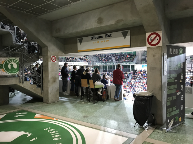 Eine Gruppe von Menschen steht vor einer sitzenden Menge in einem Stadion, mit Schildern, die "Boston Celtics vs New York Jets" anzeigen.