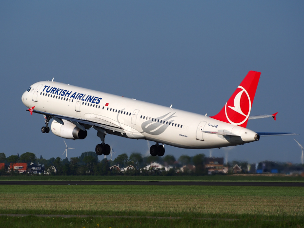 Ein Airbus A330-300 der Turkish Airlines startet am Frankfurter Flughafen, mit Gras am Boden, Gebäuden und Bäumen im Hintergrund, Windrädern in der Ferne und einem klaren blauen Himmel.